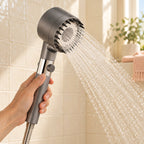 Mano sujetando la Ducha Mola gris mientras cae un chorro de lluvia en un baño de azulejos crema